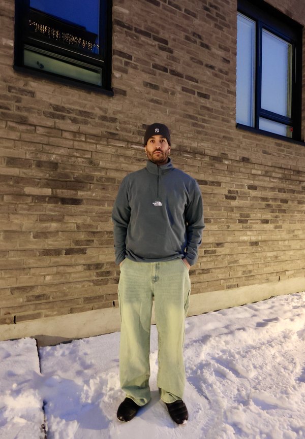 Férfi fekete sapkában, kék negyedzippes dzsekiben és világoszöld nadrágban, hóban áll egy téglából készült épület előtt alkonyatkor.