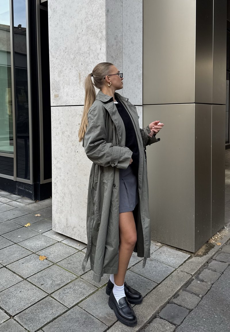 Mulher com rabo de cavalo comprido, usando óculos, casaco cinzento, topo preto, saia cinzenta, meias brancas e sapatos pretos, parada numa calçada da cidade.