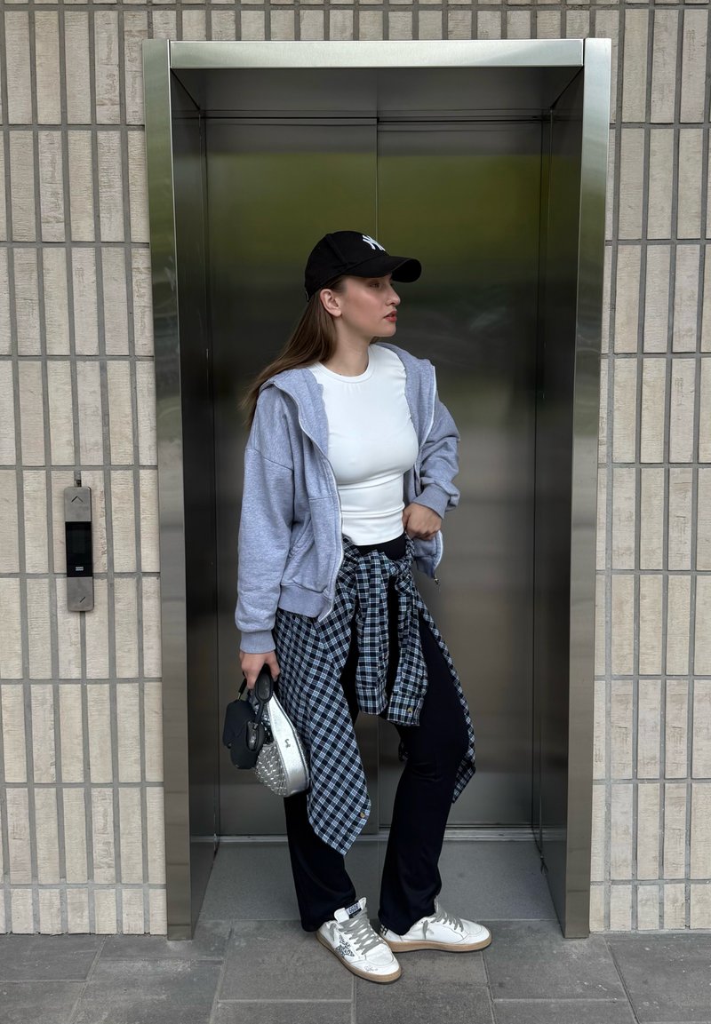 Jeune femme portant une chemise blanche, un sweat à capuche gris et une casquette noire, se tient à l'entrée d'un ascenseur en tenant deux sacs à main.