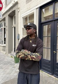 Uomo con dreadlocks che indossa occhiali da sole, un cappello nero, una maglia nera con maniche in camo e collane d'oro, in piedi su un marciapiede di città con le braccia incrociate.