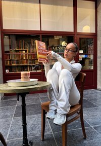 En person som sitter i en tettsittende hvit drakt, holder en bok med tittelen "Livets Spill", ved et rundt trebord med en kopp.