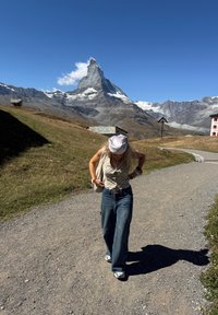 Beige rutete skjorte og lyseblå vide jeans, som går på en grusvei med fjellbakgrunn og klar blå himmel.