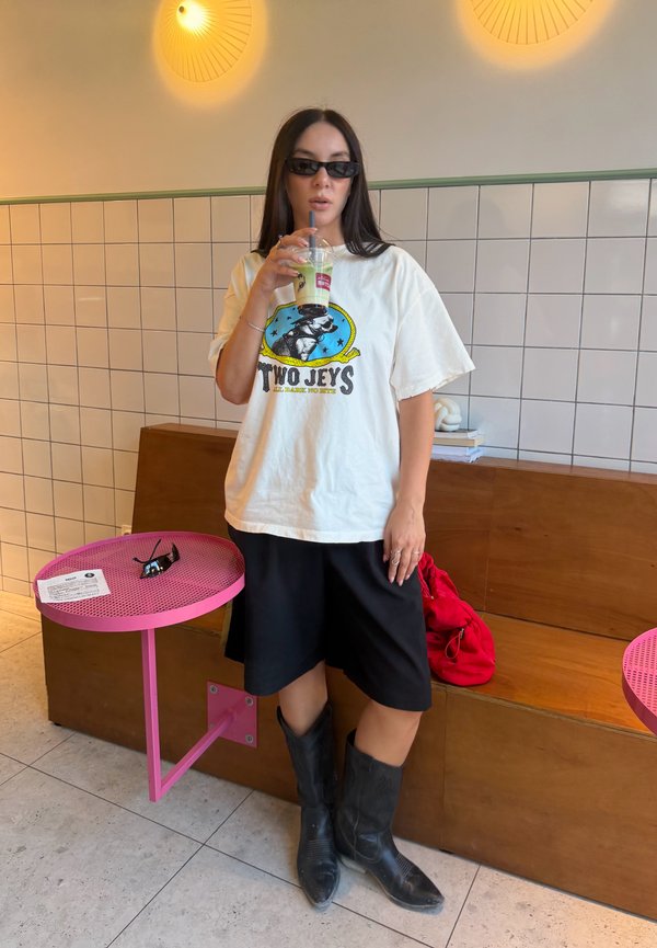 Kvinde iført solbriller, hvid grafik t-shirt, sorte shorts og støvler drikker af en kop i en flisebelagt café med lyserøde borde og træbænk.