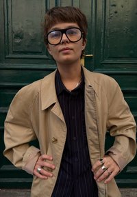Trench coat bege com grandes lapelas, usado por cima de uma camisa escura riscada. Óculos grandes pretos, anéis de prata e fundo texturizado.