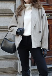 Beige dobbeltradet uldbukserjakke over en cremefarvet sweater, kombineret med sorte bukser og læderhandsker. Sort håndtaske med buet design.