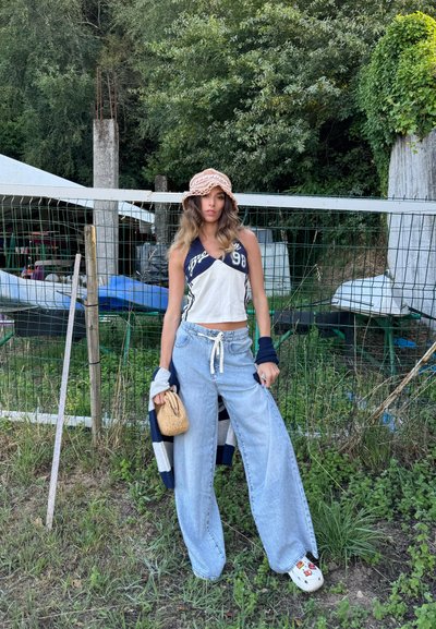 Jeans de pierna ancha de color azul claro combinados con un top recortado blanco y azul marino, llevando una bolsa de mimbre, y complementados con un sombrero tipo cubo color beige.