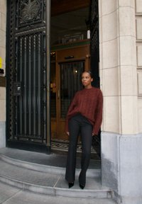 Maglione marrone a maglia con motivo a treccia, vestibilità ampia, abbinato a pantaloni neri a vita alta e a campana. L'ambiente presenta un ingresso ornato.