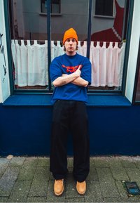 Modré oversized tričko s červeným akcentem, černé volné kalhoty, oranžová čepice a béžové boty. Pozadí tvoří okno s bílými záclonami.