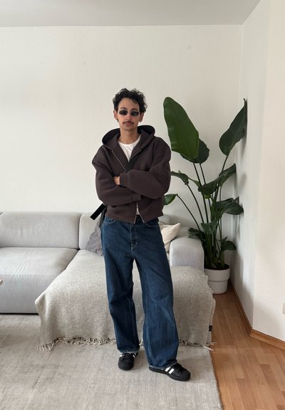Hombre con cabello rizado y gafas de sol de pie con los brazos cruzados en la sala de estar, vistiendo una sudadera oscura con capucha, jeans azules y zapatillas negras cerca de una planta verde.