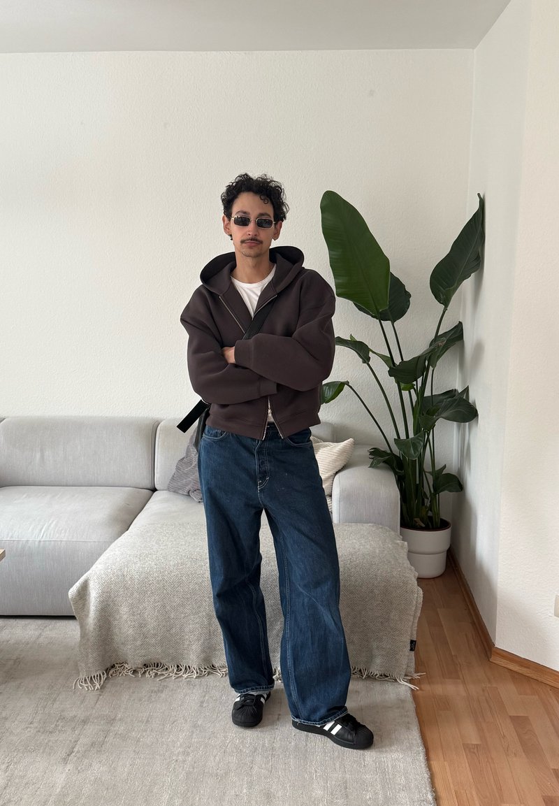 Muž s kučeravými vlasmi a slnečnými okuliarmi stojí so založenými rukami v obývačke, oblečený v tmavej mikine s kapucňou, modrých džínsoch a čiernych teniskách vedľa zelenej rastliny.