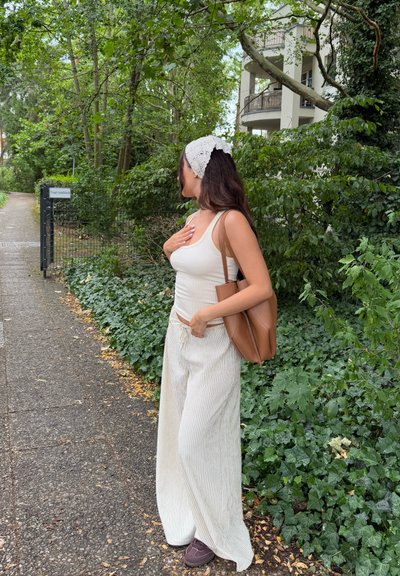 Top blanco de tirantes acanalados, pantalones de rayas anchas en beige, bolso marrón y zapatos oscuros. Cabello largo parcialmente cubierto por una diadema de encaje blanco. Fondo de follaje verde.