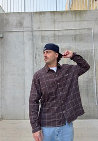 Braunes Flanellhemd mit großem grauem Karomuster, ausgestattet mit durchgehender Knopfleiste und langen Ärmeln. Kombiniert mit hellblauen Jeans.