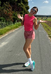 Sportolói öltözet, amely egy testhezálló rózsaszín trikót, plisszírozott rózsaszín rövidnadrágot és szürke túracipőt turquoise részletekkel tartalmaz. Nagy napszemüveg teszi teljessé a megjelenést.