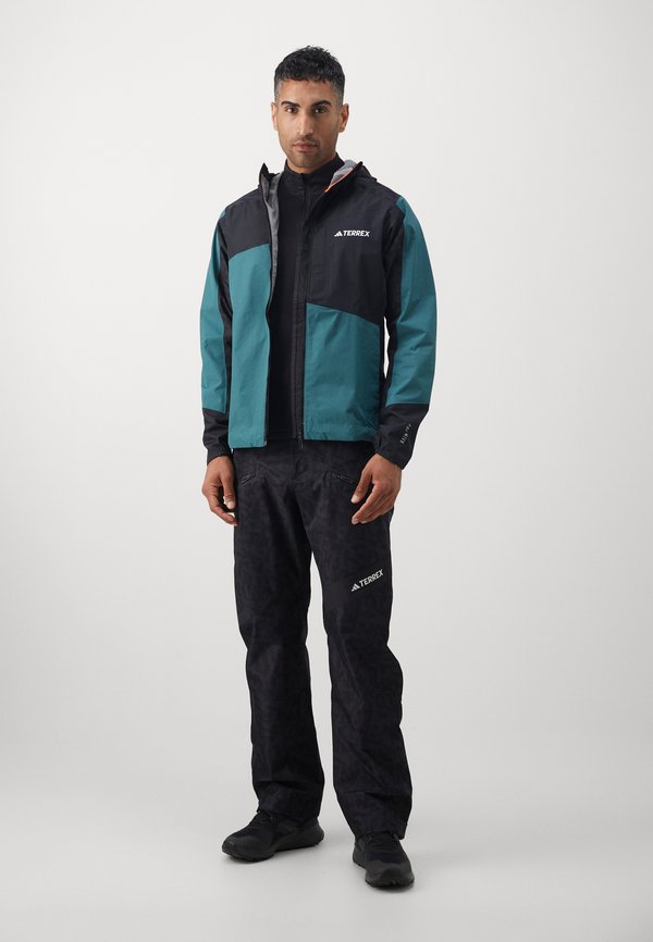 Vandeniui atsparus Terrex džemperis teal ir juodos spalvos su geometriniais raštais, derinamas su juodomis, tekstūruotomis kelnėmis, turinčiomis logotipą ir užtrauktukais kišenėse.