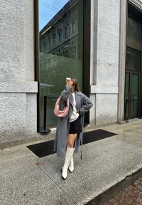 Grauer langer Mantel über einem weißen Pullover, schwarzem Minirock und weißen kniehohen Stiefeln. Hält eine Kaffeetasse und trägt eine rosa Schultertasche. Urbaner Hintergrund.
