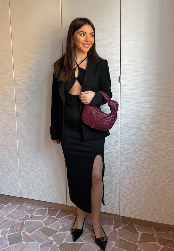 Mujer con un vestido negro con una abertura lateral y una chaqueta a juego sostiene un bolso de mano tejido burdeos, de pie sobre un suelo de mosaico.