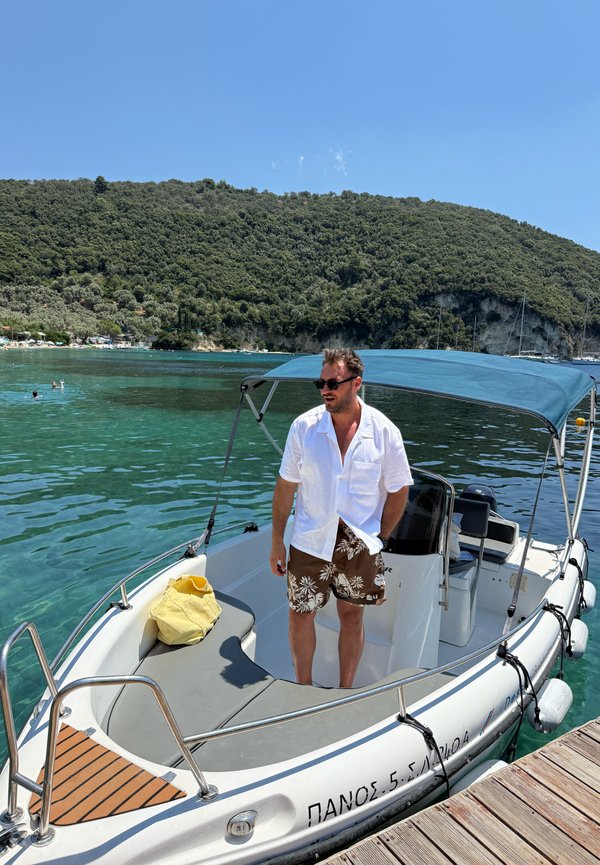Hombre con gafas de sol, camisa blanca y pantalones cortos florales, de pie en un pequeño barco atracado cerca de agua turquesa clara y colinas verdes boscosas.