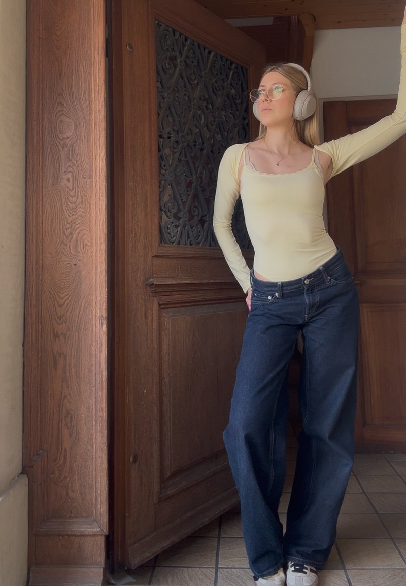 Frau mit Brille, gelbem Top, blauen Jeans und weißen Kopfhörern, die an einem hölzernen Türrahmen im Inneren lehnt.