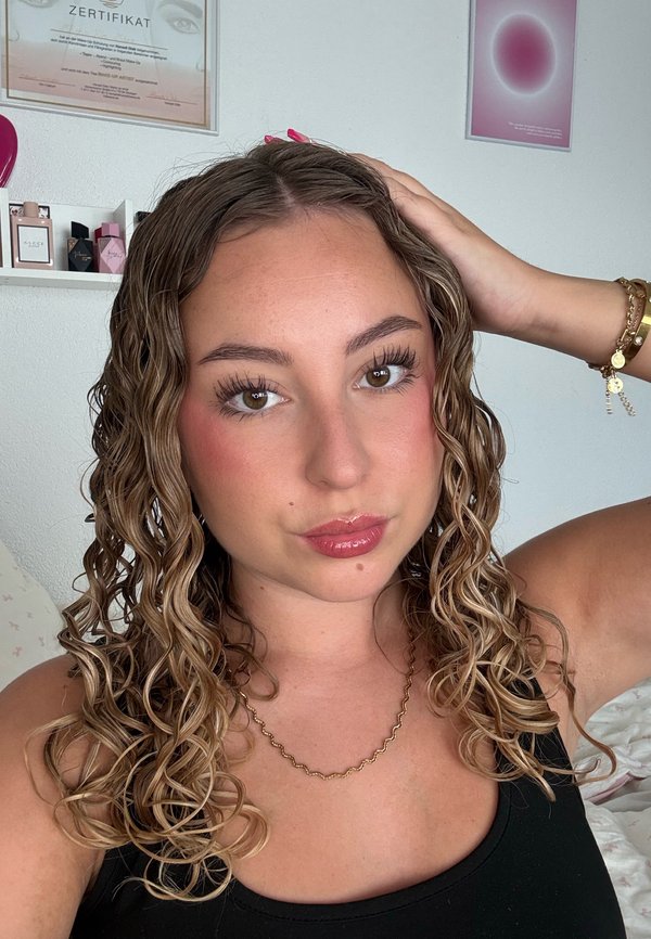 Joven mujer con cabello castaño claro ondulado tocándose la cabeza, vistiendo una camiseta negra, collar dorado y pulseras, con cuadros enmarcados en la pared detrás.