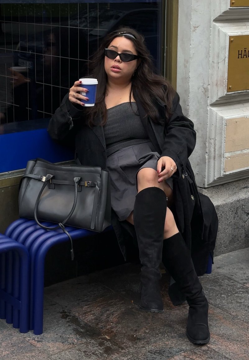 Een persoon zit op een blauwe bank, gekleed in een zwarte jas, een grijze top, een minirok en hoge zwarte laarzen, terwijl hij een koffiekop vasthoudt.
