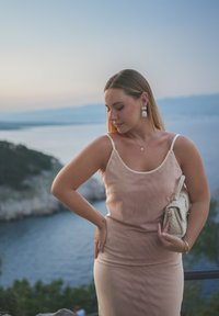 Lys rosa kjole med tynne stropper, glatt stoff og tettsittende silhuett. Tilbehør inkludert en teksturert kremfarget veske og hvite dråpeøredobber.