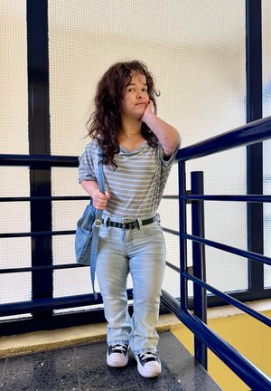 Persona joven con cabello rizado, vestida con camiseta de rayas, jeans claros y zapatillas, de pie junto a una barandilla azul dentro de un edificio, llevando una bolsa azul.
