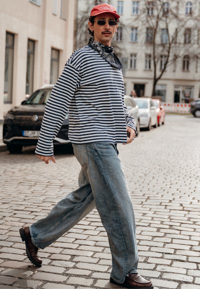 Hombre caminando por una calle empedrada, vestido con una camiseta de rayas, jeans azules, gorra roja, gafas de sol oscuras y un pañuelo negro alrededor del cuello.