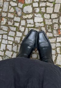 Botas de couro pretas com bico quadrado, apresentando uma textura lisa e um leve brilho, posicionadas sobre uma superfície de calçada de pedra.