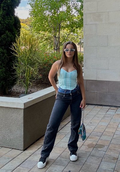 Top de corsé color aguamarina con costuras verticales, jeans negros de tiro alto, zapatillas blancas y un bolso mini con estampado. Entorno urbano al aire libre.