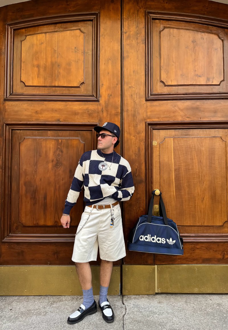 Pull en tricot à motif à carreaux bleu marine et crème, shorts beiges, chaussures noires, chaussettes grises. Sac Adidas reposant contre une porte en bois.