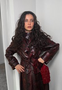 Trench coat em padrão de pele de cobra marrom-escuro, com uma cintura cingida por cinto e botões grandes, acompanhado de uma bolsa de pérolas vermelha. Textura lisa e brilhante.