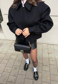 Cazadora bomber negra de mezcla de lana con cuello alto, combinada con una falda mini de cuero negra, bolso negro y zapatos negros brillantes con calcetines blancos.