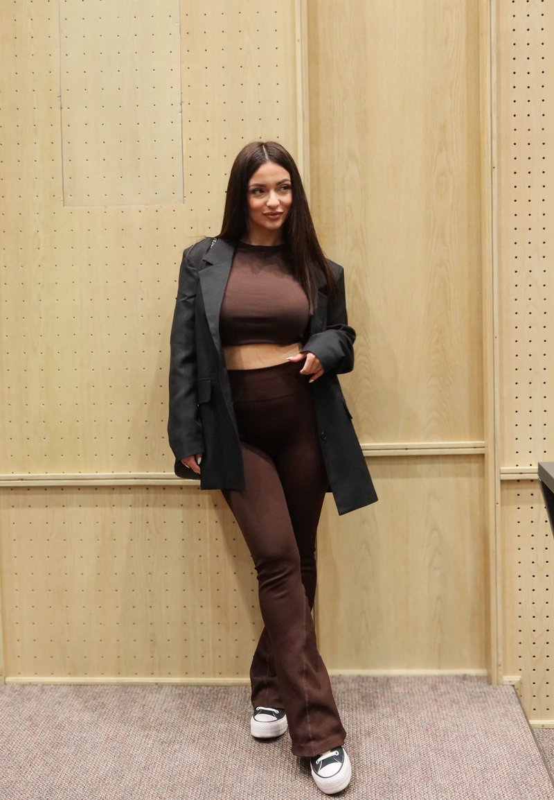 Donna con capelli lunghi e scuri, indossa un top corto marrone e pantaloni abbinati, con un blazer nero e scarpe da ginnastica bianche, in piedi davanti a una parete in legno chiaro.