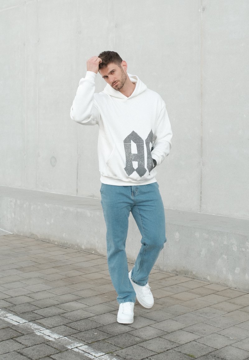 Homme portant un sweat à capuche blanc et un jean bleu se tenant sur une surface pavée, se grattant la tête devant un mur en béton uni.