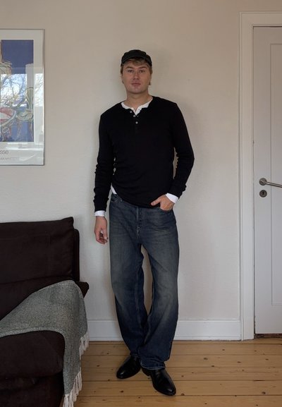 Hombre vestido con camisa negra de manga larga, pantalones vaqueros y una gorra negra, de pie en el interior cerca de un sofá marrón con una manta gris y una puerta blanca cerrada.