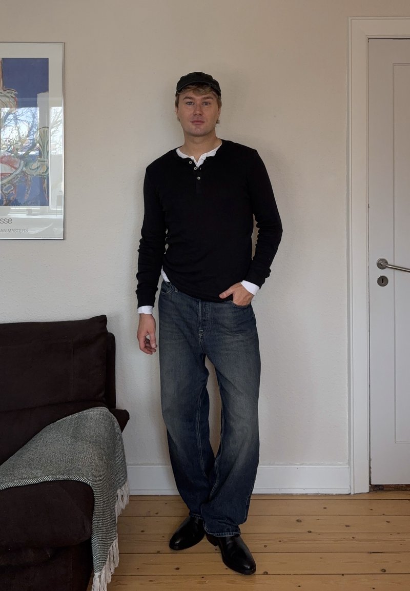 Homme portant une chemise noire à manches longues, un jean et une casquette noire, debout à l'intérieur près d'un canapé marron avec une couverture grise et une porte blanche fermée.