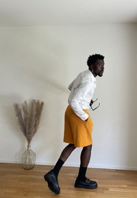 Man in white shirt, orange shorts, and black shoes with socks holding glasses, leaning forward indoors near a vase with pampas grass.