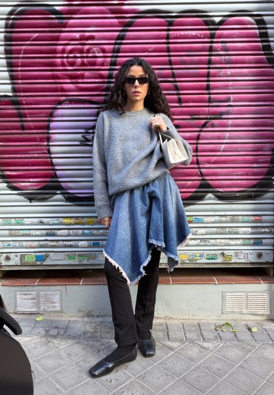 Suéter gris, falda vaquera con dobladillo desflecado, pantalones negros y zapatos negros. La modelo sostiene un pequeño bolso beige frente a un fondo de graffiti rosa.