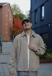 Beige suede jacket with a collar, front zip, and buttons; layered over a patterned shirt. Casual grey trousers and a green cap.