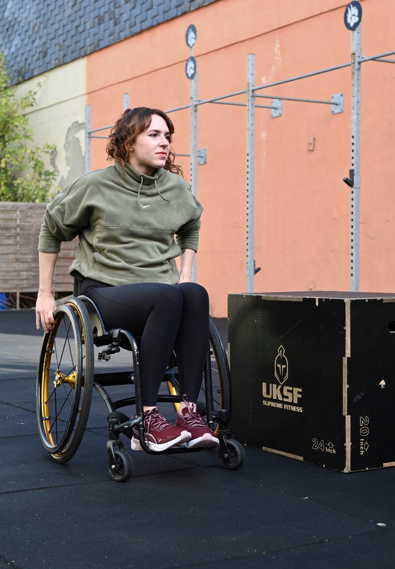Ein Rollstuhl mit goldenen Akzenten unterstützt eine Person in einem grünen Sweatshirt und schwarzen Leggings neben einer schwarzen Fitnessbox auf einer Gummimatte.