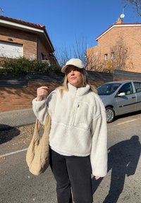 Jeune femme portant une veste en polaire blanche et une casquette beige, debout dans la rue tenant un sac matelassé beige, avec une voiture garée et des maisons en briques en arrière-plan.