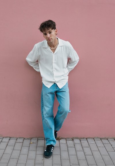 Camisa a rayas blancas, jeans de mezclilla azul claro y zapatillas negras. Se lleva un collar de perlas. El fondo presenta una pared rosa y un pavimento gris.