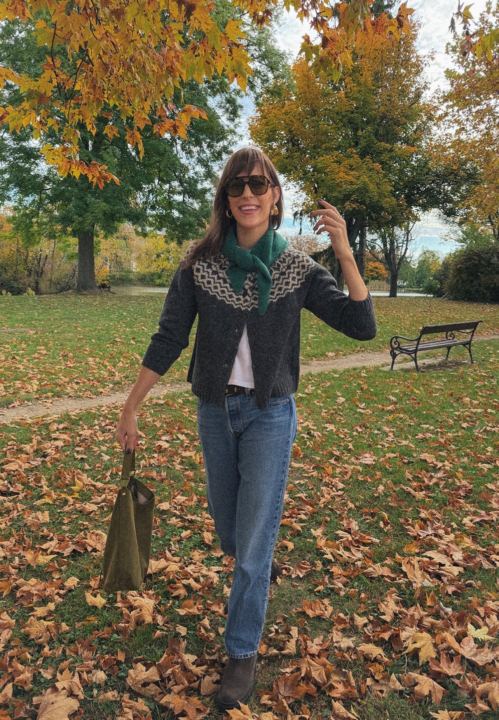 Cardigan lavorato a maglia con una base grigio scuro e un motivo sulla spalla di colore chiaro, abbinato a jeans blu e stivaletti marroni, mentre tiene una borsa verde.