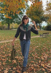 Cardigan lavorato a maglia con una base grigio scuro e un motivo sulla spalla di colore chiaro, abbinato a jeans blu e stivaletti marroni, mentre tiene una borsa verde.
