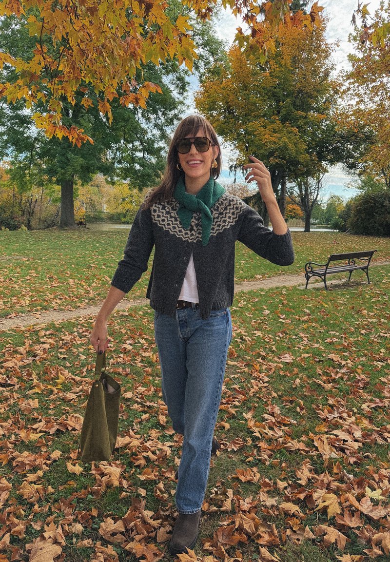 Strikket cardigan med en mørkegrå base og mønster i lyse farger på bærestykket, kombinert med blå jeans og brune ankelboots, og holder en grønn veske.