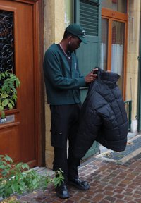 Giubbotto invernale nero tenuto da una persona che indossa un cardigan verde e pantaloni cargo neri. La scena presenta un pavimento in mattoni e una porta di legno.