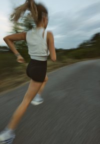 Løper i hvit tank topp og svarte shorts, iført blå løpesko. Bakgrunnen er uskarp, noe som formidler bevegelse langs en sti.