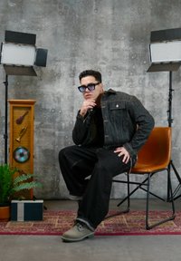 Homme en veste et pantalon en jean foncé, portant des lunettes teintées de bleu, assis sur une chaise marron entre deux lampes de studio, devant un mur en béton.