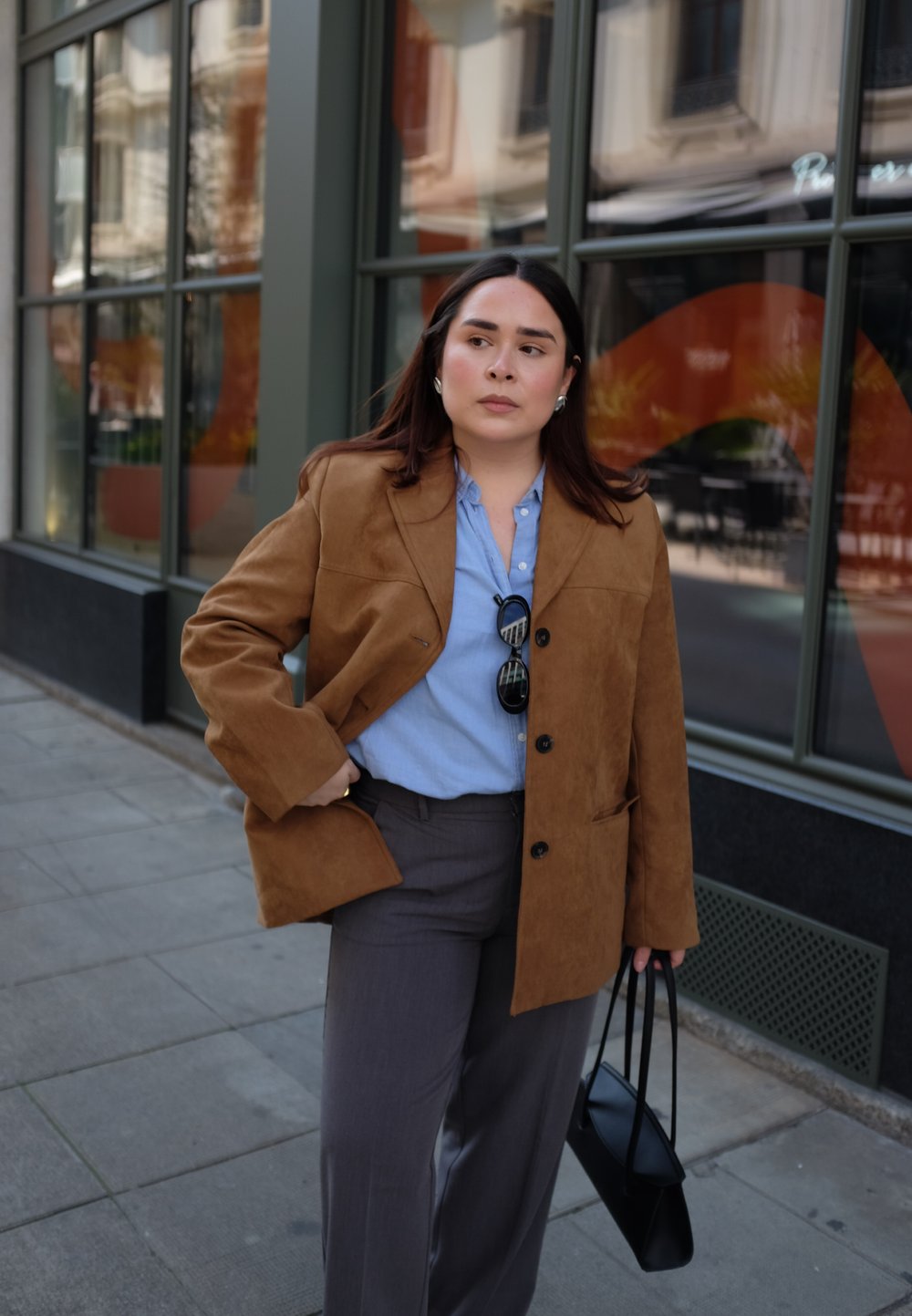Bruine suède blazer over een lichtblauw shirt, gecombineerd met grijze broek. Houdt een zwarte handtas vast. Stedelijke omgeving met grote ramen op de achtergrond.