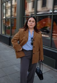 Bruine suède blazer over een lichtblauw shirt, gecombineerd met grijze broek. Houdt een zwarte handtas vast. Stedelijke omgeving met grote ramen op de achtergrond.